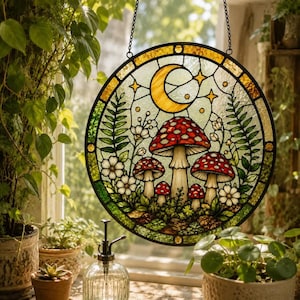 Op de afbeelding: Een ronde glas-in-lood zonnevanger met een ontwerp van rode en witte paddenstoelen, omringd door groene varens en kleine witte bloemen. Een gele maansikkel en sterren zijn afgebeeld in het bovenste gedeelte tegen een lichte achtergrond.