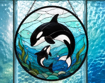 May include: A circular stained glass suncatcher featuring a black and white orca and a smaller calf swimming in a blue ocean scene with green seaweed. The piece is framed in black metal and hangs from a black chain against a window background.