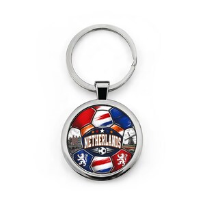 May include: A circular silver keychain featuring a soccer ball graphic with red, white, and blue panels. The design includes the text NETHERLANDS, a windmill, a building, and lion emblems. The metal ring is attached to the top of the round pendant.