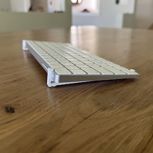 May include: A silver and white wireless keyboard is elevated at an angle by two white plastic stands attached to its sides. The keyboard features a low-profile design with white square keys and a metallic silver frame, resting on a wooden surface.