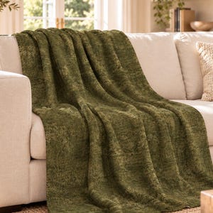 May include: A textured olive green throw blanket draped over a light beige sofa. The blanket features a dense, plush pile with subtle tonal variations in the green fabric, creating a soft, uneven surface appearance across the entire piece.