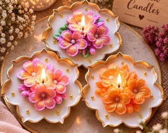 May include: Three flower-shaped candles on white scalloped plates with gold rims. Each candle features a cluster of wax flowers in pink, orange, and yellow with gold accents and small white beads. The candles are lit, and a wooden sign reads Scented Candles.