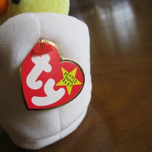 Peut inclure: Un jouet en peluche jaune repr&eacute;sentant un poussin sortant d'une coquille d'&oelig;uf en tissu blanc. Une &eacute;tiquette rouge en forme de c&oelig;ur avec les lettres ty en blanc et une &eacute;toile jaune portant le texte Beanie Original Baby est fix&eacute;e sur le c&ocirc;t&eacute;.