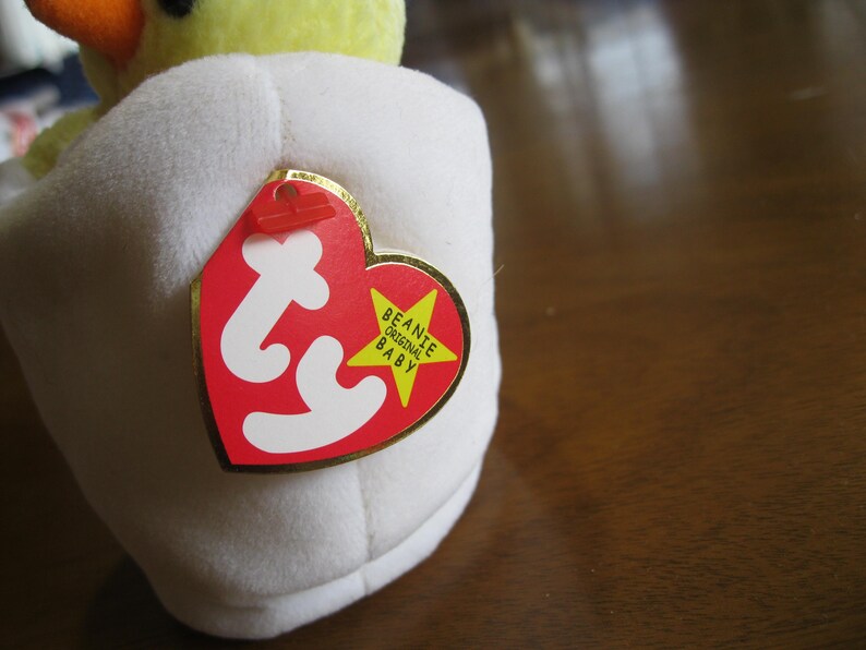 Peut inclure: Un jouet en peluche jaune repr&eacute;sentant un poussin sortant d'une coquille d'&oelig;uf en tissu blanc. Une &eacute;tiquette rouge en forme de c&oelig;ur avec les lettres ty en blanc et une &eacute;toile jaune portant le texte Beanie Original Baby est fix&eacute;e sur le c&ocirc;t&eacute;.
