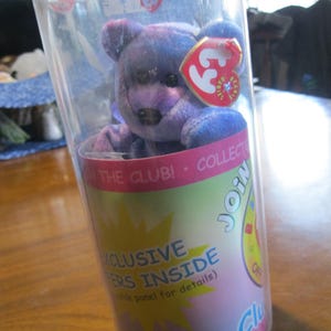 May include: A purple and blue tie-dye plush bear inside a clear plastic cylindrical container. The container features a pink and yellow label with the text JOIN THE CLUB, EXCLUSIVE OFFERS INSIDE, and a red heart-shaped Ty Beanie Babies tag.