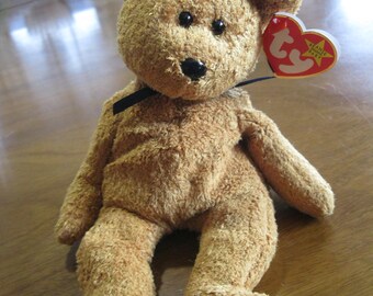 May include: A tan plush teddy bear with a textured, fuzzy surface, black plastic eyes, and a black plastic nose. A thin black ribbon is tied around its neck. A heart-shaped red and yellow tag with the text "ty" and "Beanie Baby" is attached.
