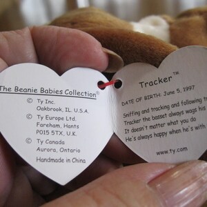 May include: A heart-shaped white paper tag from The Beanie Babies Collection attached to a brown plush toy. The tag features the name Tracker, a birth date of June 5, 1997, and text detailing the manufacturer locations in the U.S., U.K., and Canada.
