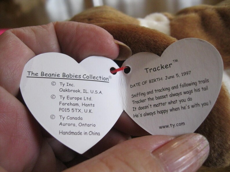 May include: A heart-shaped white paper tag from The Beanie Babies Collection attached to a brown plush toy. The tag features the name Tracker, a birth date of June 5, 1997, and text detailing the manufacturer locations in the U.S., U.K., and Canada.