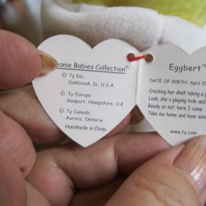 Peut inclure: Une &eacute;tiquette blanche en forme de c&oelig;ur de la collection Ty Beanie Babies pour Eggbert. Le c&ocirc;t&eacute; gauche indique les sites de Ty Inc. aux &Eacute;tats-Unis, au Royaume-Uni et au Canada, et mentionne Handmade in China. Le c&ocirc;t&eacute; droit affiche le nom Eggbert, la date de naissance 10 avril 1998 et un po&egrave;me.