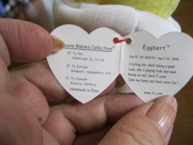 Peut inclure: Une &eacute;tiquette blanche en forme de c&oelig;ur de la collection Ty Beanie Babies pour Eggbert. Le c&ocirc;t&eacute; gauche indique les sites de Ty Inc. aux &Eacute;tats-Unis, au Royaume-Uni et au Canada, et mentionne Handmade in China. Le c&ocirc;t&eacute; droit affiche le nom Eggbert, la date de naissance 10 avril 1998 et un po&egrave;me.