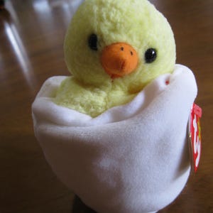 Peut inclure: Une peluche repr&eacute;sentant un petit poussin jaune duveteux avec des yeux noirs et un bec orange, nich&eacute; dans une coquille en forme d'&oelig;uf en tissu blanc doux. Une &eacute;tiquette rouge en forme de c&oelig;ur avec les lettres ty est fix&eacute;e sur le c&ocirc;t&eacute;.