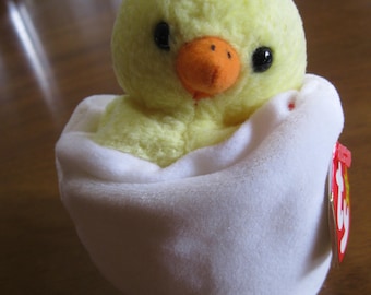 May include: A plush toy featuring a small, fuzzy yellow chick with black eyes and an orange beak, nestled inside a soft white egg-shaped shell. A red heart-shaped tag with the letters ty is attached to the side of the white plush egg shell.