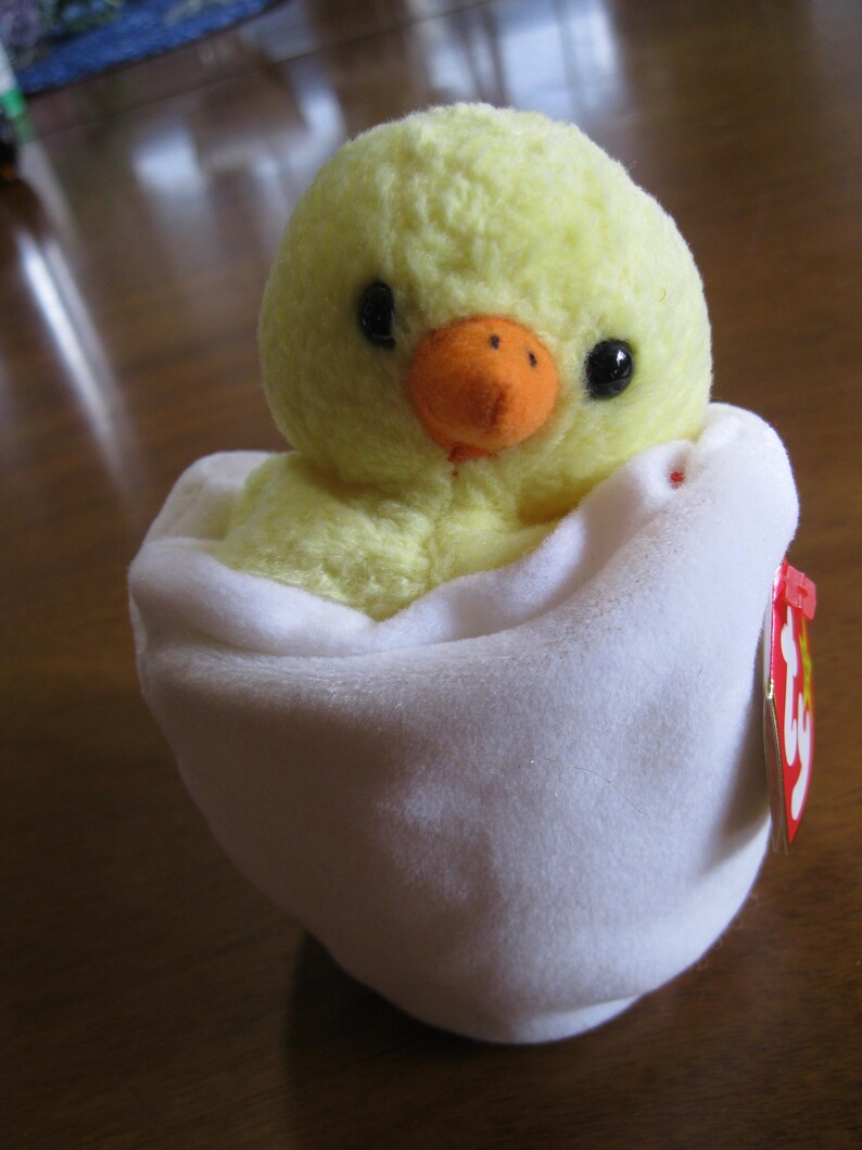 Peut inclure: Une peluche repr&eacute;sentant un petit poussin jaune duveteux avec des yeux noirs et un bec orange, nich&eacute; dans une coquille en forme d'&oelig;uf en tissu blanc doux. Une &eacute;tiquette rouge en forme de c&oelig;ur avec les lettres ty est fix&eacute;e sur le c&ocirc;t&eacute;.