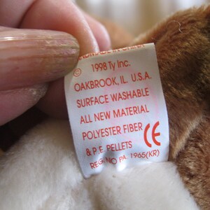 May include: A white fabric tag attached to a brown plush toy. The tag features red text reading 1998 Ty Inc. OAKBROOK, IL. U.S.A. SURFACE WASHABLE ALL NEW MATERIAL POLYESTER FIBRE & P.E. PELLETS REG. NO PA. 1965(KR) and a red CE symbol.