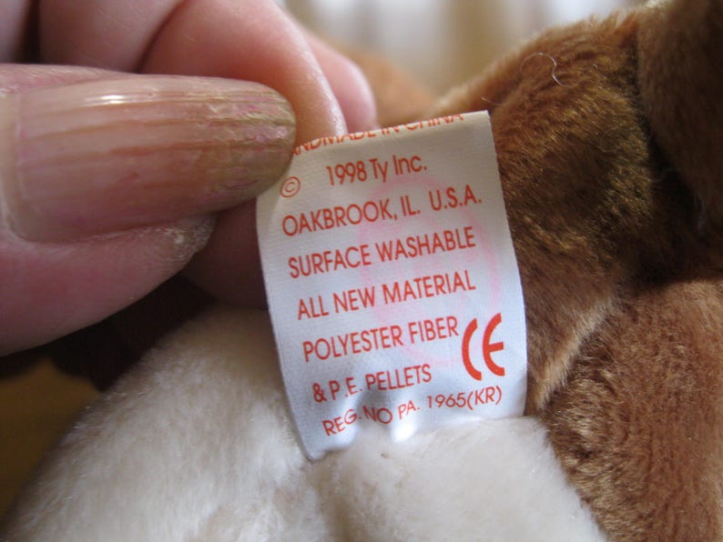 May include: A white fabric tag attached to a brown plush toy. The tag features red text reading 1998 Ty Inc. OAKBROOK, IL. U.S.A. SURFACE WASHABLE ALL NEW MATERIAL POLYESTER FIBRE & P.E. PELLETS REG. NO PA. 1965(KR) and a red CE symbol.
