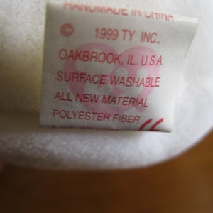 Peut inclure: Une &eacute;tiquette en tissu blanc provenant d'une peluche avec du texte rouge indiquant : HANDMADE IN CHINA, 1999 TY INC., OAKBROOK, IL. U.S.A., SURFACE WASHABLE, ALL NEW MATERIAL, POLYESTER FIBER. Un logo rose en forme de c&oelig;ur est visible.