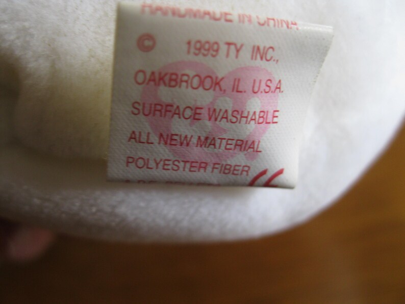 Peut inclure: Une &eacute;tiquette en tissu blanc provenant d'une peluche avec du texte rouge indiquant : HANDMADE IN CHINA, 1999 TY INC., OAKBROOK, IL. U.S.A., SURFACE WASHABLE, ALL NEW MATERIAL, POLYESTER FIBER. Un logo rose en forme de c&oelig;ur est visible.