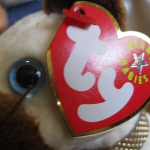 May include: A plush toy featuring a cream and brown fur texture with a single visible dark eye. A heart-shaped red tag with white 'ty' lettering and the words 'BEANIE BABIES' in yellow text is attached to the toy with a red plastic fastener.