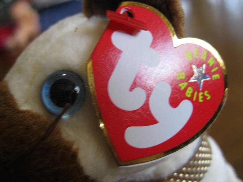 May include: A plush toy featuring a cream and brown fur texture with a single visible dark eye. A heart-shaped red tag with white 'ty' lettering and the words 'BEANIE BABIES' in yellow text is attached to the toy with a red plastic fastener.