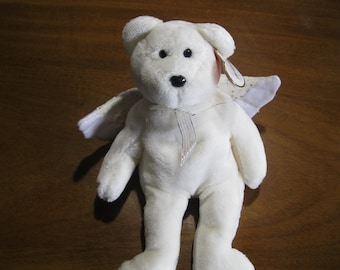Peut inclure: Un ours en peluche blanc avec une fourrure douce, des yeux en plastique noir et un nez noir. L'ours porte un ruban transparent noué autour du cou et une paire d'ailes en tissu blanc fixées dans le dos. Une étiquette en forme de cœur est attachée à l'oreille.