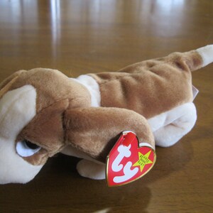 Puede incluir: Un perro de peluche con pelaje marrón y blanco, orejas largas y caídas, y una nariz negra. El juguete tiene una etiqueta roja y amarilla en forma de corazón con el texto ty Beanie Original Baby impreso en letras blancas y negras.