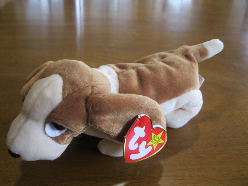May include: A plush toy dog with brown and white fur, long floppy ears, and a black nose. The toy features a heart-shaped red and yellow tag attached to its ear with the text ty Beanie Original Baby printed in white and black lettering.