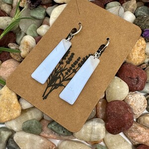 May include: A pair of long, rectangular, light blue earrings with a smooth finish, attached to a brown cardboard display card featuring a black line drawing of lavender sprigs and silver-toned metal earring hooks for pierced ears.
