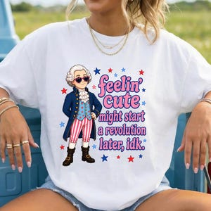 May include: A white short-sleeve t-shirt featuring a cartoon illustration of a historical figure wearing a blue coat, red and white striped pants, and heart-shaped sunglasses. The text reads, "feelin' cute might start a revolution later, idk."