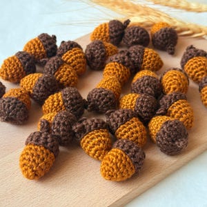 Può includere: Una collezione di piccole ghiande realizzate a mano all'uncinetto, con frutti giallo oro e cupole testurizzate marrone scuro. Questi ornamenti tessili individuali sono sparsi su una superficie in legno chiaro, mostrando dettagliate cuciture.