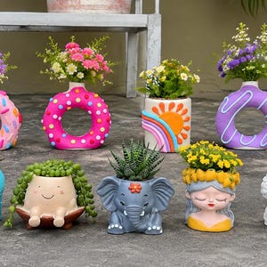 May include: A collection of thirteen small ceramic planters in various shapes including doughnuts, animals, and human faces. The planters feature colourful designs like polka dots, rainbows, and cacti, each holding a small succulent or floral arrangement.