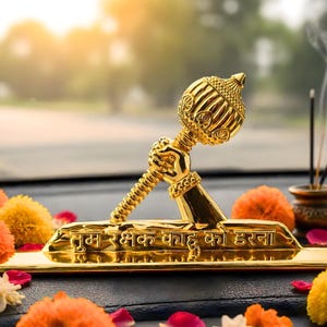 Puede incluir: Una figura decorativa de color dorado que representa una mano sosteniendo una maza, montada sobre una base rectangular. La base muestra el texto en hindi "तुम रक्षक काहू को डरना" en letras doradas en relieve. El objeto tiene un acabado metálico pulido.