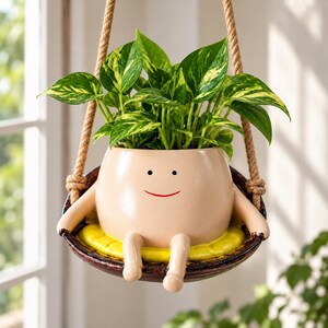 Puede incluir: Una maceta de cerámica beige con forma de figura sentada con una sonrisa roja y ojos negros, sobre un columpio circular marrón oscuro con un cojín amarillo. Una planta pothos variegada verde y amarilla crece en la maceta suspendida por cuerdas.