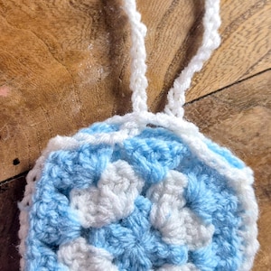 May include: A circular crochet ornament featuring a repeating floral pattern in light blue and white yarn. The design consists of a central white flower motif surrounded by light blue petals, finished with a white border and a matching hanging loop.