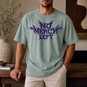 May include: A sage green short-sleeved t-shirt featuring a dark purple graphic print on the chest. The text reads NO MERCY LEFT in a sharp, jagged, stylized font. The garment has a crew neckline and a relaxed fit, constructed from cotton fabric.