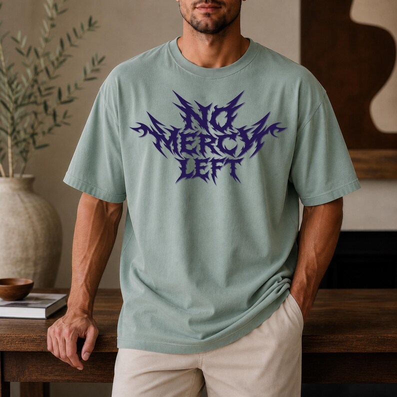 May include: A sage green short-sleeved t-shirt featuring a dark purple graphic print on the chest. The text reads NO MERCY LEFT in a sharp, jagged, stylized font. The garment has a crew neckline and a relaxed fit, constructed from cotton fabric.