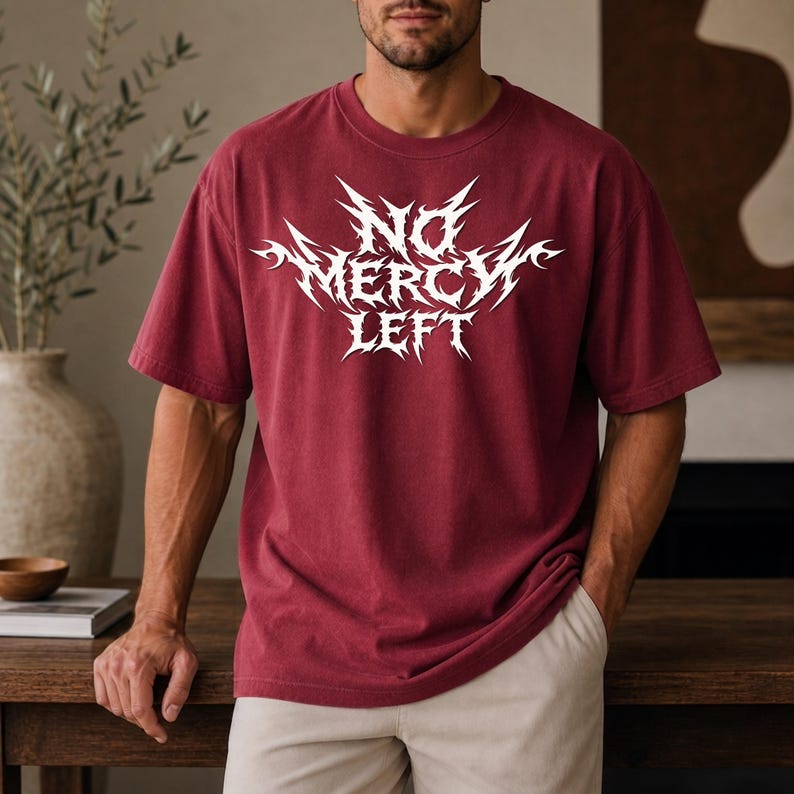 May include: A maroon short-sleeved t-shirt featuring a white graphic print on the chest with the text "NO MERCY LEFT" in a stylized, sharp, angular font. The garment has a crew neckline and a relaxed fit, constructed from a solid-colored fabric.