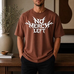 May include: A brown short-sleeved t-shirt featuring a white graphic print on the chest with the text NO MERCY LEFT in a sharp, jagged, stylized font. The garment has a crew neckline and a relaxed fit, constructed from a solid-colored fabric.