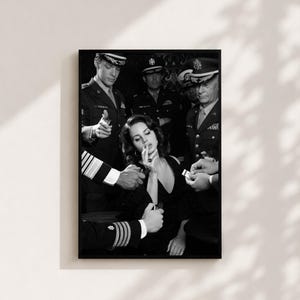 May include: A black and white framed art print depicting a woman in a dark dress surrounded by several individuals in military uniforms. The figures are shown lighting a cigarette for the woman, with focus on the contrast of the dark attire.