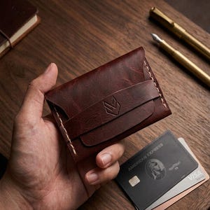 May include: A dark brown leather card holder featuring a textured surface, a horizontal strap across the front with a stamped geometric logo, and visible light-colored stitching along the edges. The wallet is rectangular with rounded corners.