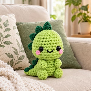 May include: A light green crochet dinosaur plush toy with a large round head, dark green spikes along its back, black button eyes, and small pink stitched cheeks. The stuffed creature has a smiling expression and a textured yarn finish.