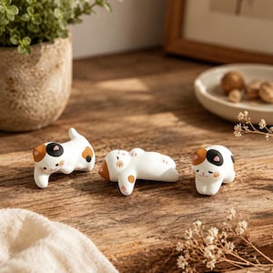 May include: Three small ceramic cat figurines in white with black and orange patches. Each cat is depicted in a different resting pose, featuring painted facial details including eyes, noses, and pink ears. The figures have smooth surfaces.