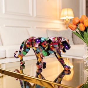 Peut inclure: Une figurine décorative de panthère arborant un motif graffiti multicolore et vibrant avec des éclaboussures de rouge, bleu, jaune et noir. La sculpture représente l'animal dans une posture de rôdeur, la gueule ouverte, révélant une peinture détaillée.