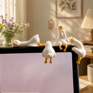 Könnte beinhalten: Ein Set aus vier kleinen weißen Gänsefiguren mit gelben Schnäbeln und Füßen, die auf dem oberen Rand eines schwarzen Monitorrahmens sitzen. Eine Gans liegt flach, eine steht, eine sitzt aufrecht mit einem Flügel am Gesicht, eine lehnt sich über.