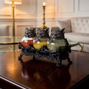 May include: A set of three clear glass jars with black cat-shaped lids featuring red eyes, held in a black ornate stand with scrollwork details. The jars contain red, yellow, and green spices, resting on a dark wooden surface in a room setting.