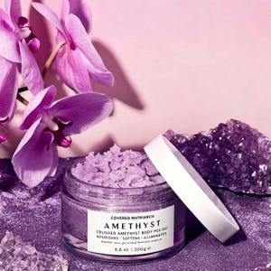 May include: A clear glass jar of light purple body polish with a white lid. The label reads COVERED MATRIARCH AMETHYST CRUSHED AMETHYST BODY POLISH NOURISHES - SOFTENS - ILLUMINATES Awaken your grounded feminine essence 6.6 oz | 200g e.