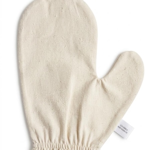 May include: An off-white, natural-colored fabric mitt with a thumb section and an elasticized wrist cuff. A small white fabric tag is attached to the side, featuring the black printed text "COVERED MATRIARCH" in a clean, sans-serif font.