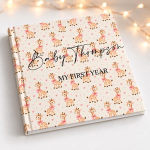 May include: A square photo album with a cream-coloured cover featuring a repeating pattern of watercolour giraffes wearing pink bows and small pink hearts. The text Baby Thompson and MY FIRST YEAR is printed in black on the front cover.