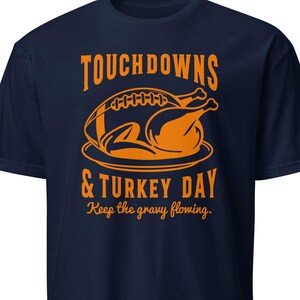 May include: A navy blue short-sleeve t-shirt featuring an orange graphic of a football and a roasted turkey on a platter. The text reads TOUCHDOWNS & TURKEY DAY and Keep the gravy flowing in a bold, stylized orange font centred on the chest.