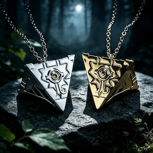 May include: Two pyramid-shaped pendants on metal chains, one in a polished silver tone and one in a polished gold tone. Each pendant features an engraved eye symbol on the front face, with a small circular accent at the center of the eye design.