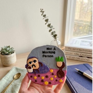 Op de afbeelding: Een ronde kleiplaque met een grijze achtergrond en de tekst Not A Morning Person. Een cartoon toast-figuurtje met een verdrietig gezicht houdt een mok vast terwijl het op een paarse wolk rust boven een roze oppervlak met gele en bruine stippen.